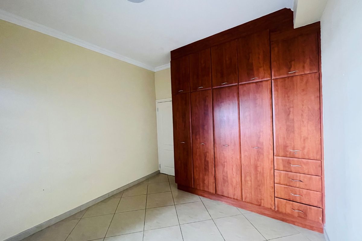 A bright, empty bedroom featuring extensive floor-to-ceiling built-in wooden cupboards and neutral tiled flooring.