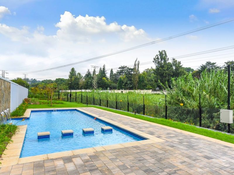 A well-maintained outdoor pool area featuring a stone-clad water feature, paved decking, and landscaped lawn, set within a secure apartment complex.