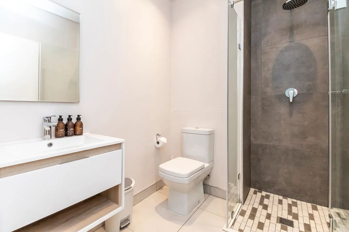 Modern bathroom featuring a sleek vanity, wall-mounted mirror, white toilet, and a spacious walk-in shower with dark tiled walls and mosaic floor detailing.