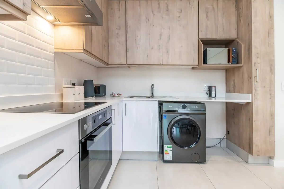 Modern kitchen featuring light wood-tone cabinetry, white subway tile backsplash, stone countertops, integrated oven, electric hob, and a dedicated space for laundry appliances.