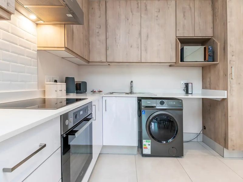 Modern kitchen featuring light wood-tone cabinetry, white subway tile backsplash, stone countertops, integrated oven, electric hob, and a dedicated space for laundry appliances.