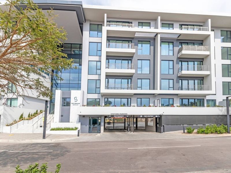 Modern apartment building exterior featuring a glass facade, multiple balconies with metal railings, and a dedicated underground visitors' parking entrance.
