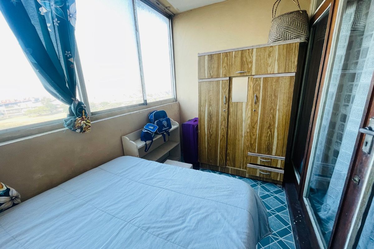 A compact bedroom featuring large windows providing natural light and tiled flooring, currently requiring cosmetic updates to the wall finishes and window frames.