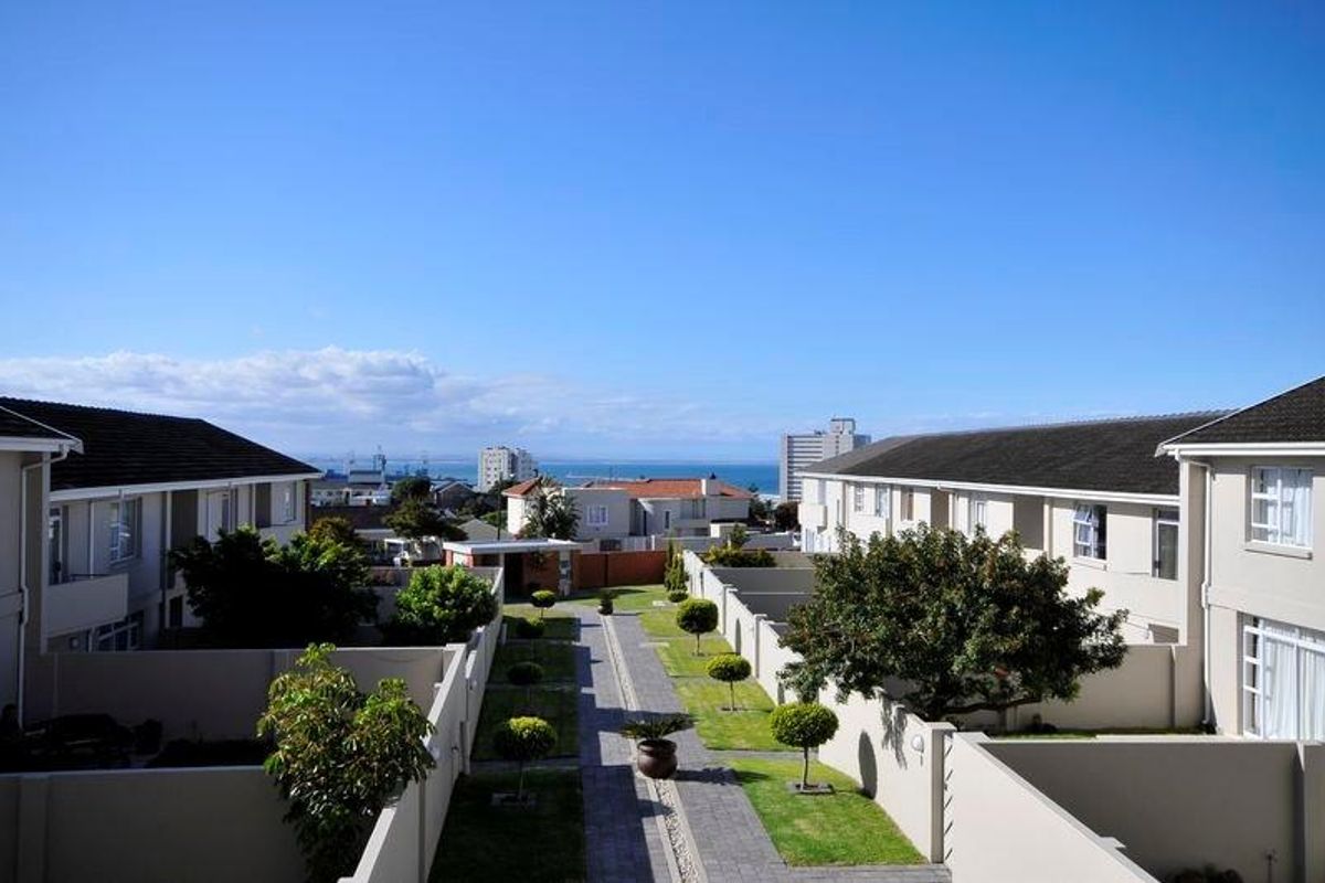 An elevated view of the rear communal garden area between apartment blocks, featuring paved pathways, manicured trees, and boundary walls with a distant sea view.