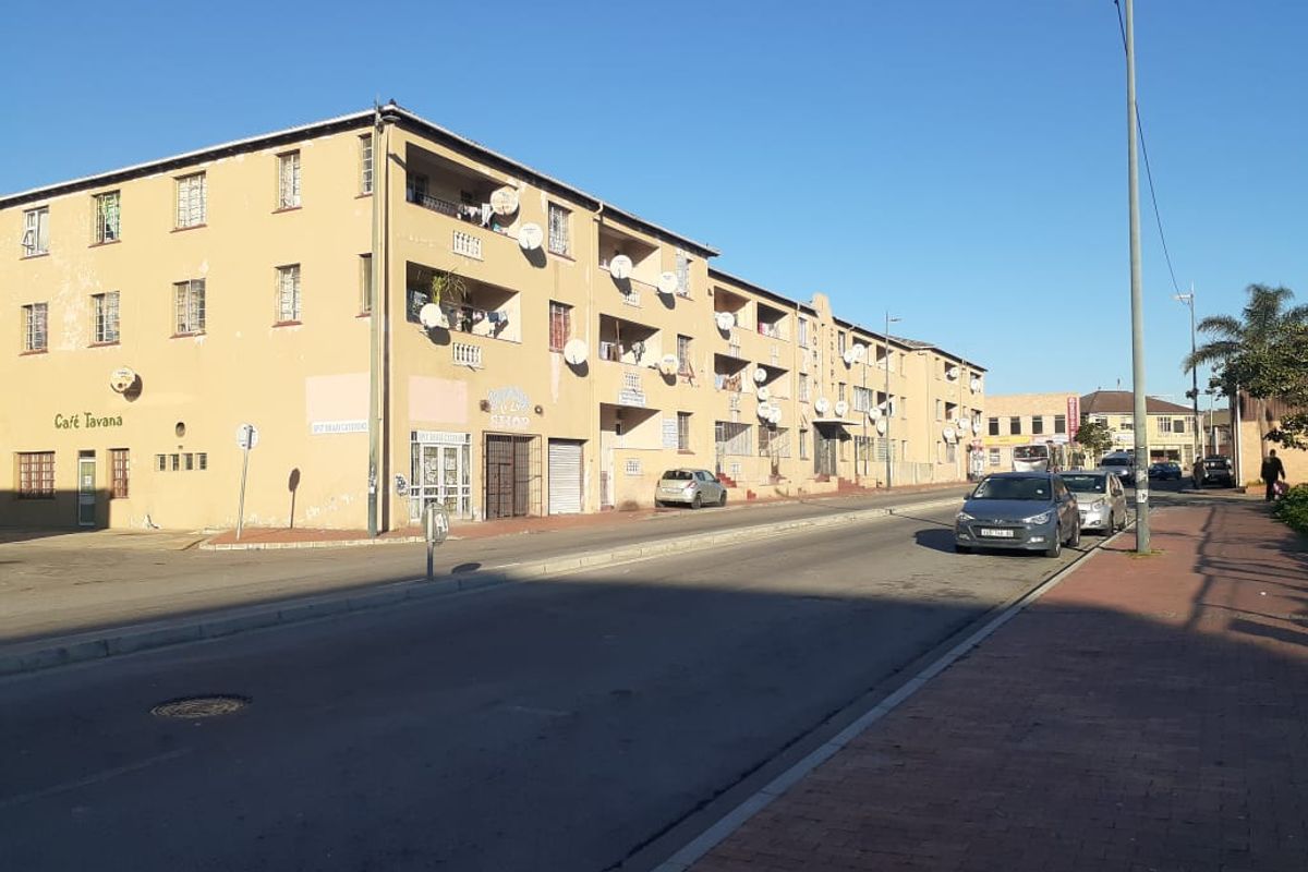 Street view of a multi-story apartment building featuring balconies and ground-level commercial space, showing signs of exterior wear.