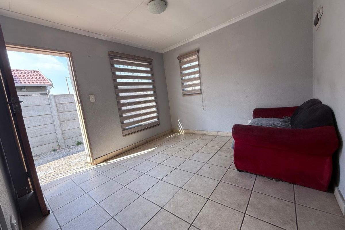 A simple living room featuring neutral ceramic floor tiles, roller blinds, and direct access to the exterior through a standard door.