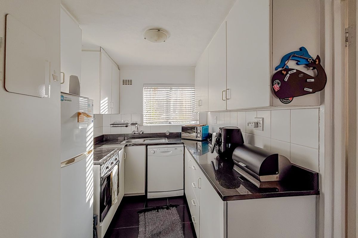 A functional galley-style kitchen featuring white cabinetry, dark stone countertops, tiled backsplash, and integrated cooking appliances with natural light from a window.