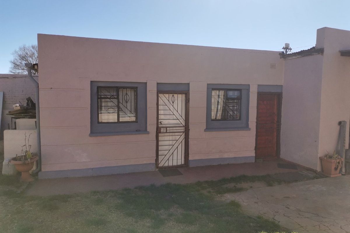 Rear exterior view of a single-story residential building featuring plastered walls, security gates, and burglar bars with an adjacent concrete paved area.