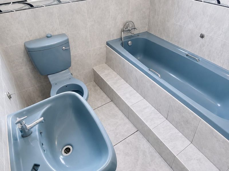 A clean, functional bathroom featuring a blue suite, including a built-in bathtub, pedestal sink, and toilet, complemented by neutral wall and floor tiling.
