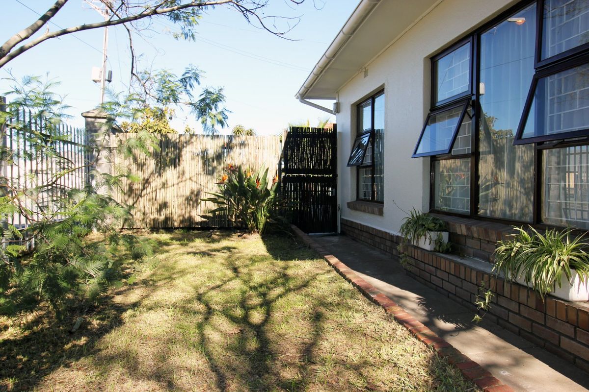 A view of the rear garden featuring a paved walkway, brick foundation detailing, and secure casement windows along the side of the house.