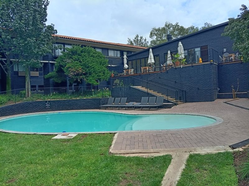 Communal swimming pool area featuring a paved deck, raised patio with metal railings, and landscaped garden surroundings within the apartment complex.