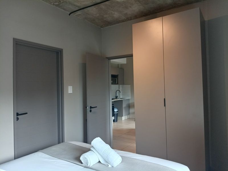A modern bedroom featuring an exposed concrete ceiling, built-in wardrobe, and direct access to the kitchen area.