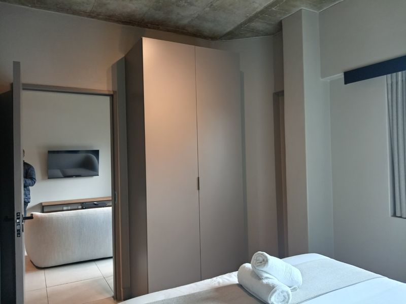 A modern bedroom featuring a built-in wardrobe, exposed concrete ceiling, and tiled flooring, with an adjacent view into the living area.