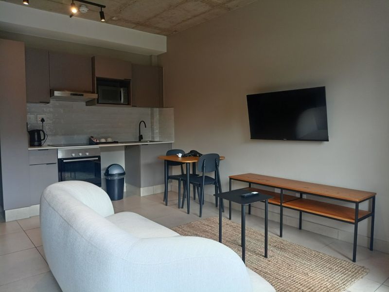 Modern open-plan apartment featuring a compact kitchen with built-in appliances, tiled flooring, and an exposed concrete ceiling in the living area.