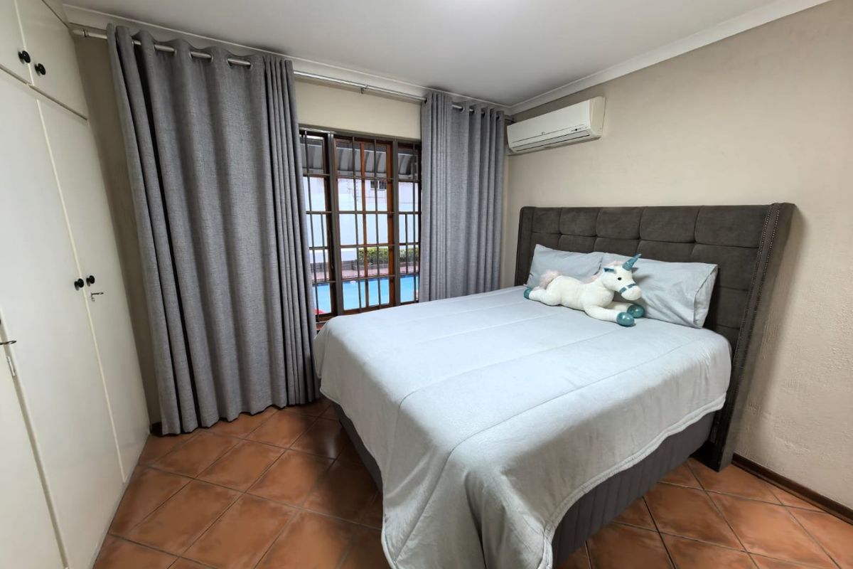 A well-maintained bedroom featuring tiled flooring, built-in cupboards, and a wall-mounted air conditioning unit, with views overlooking the pool area.
