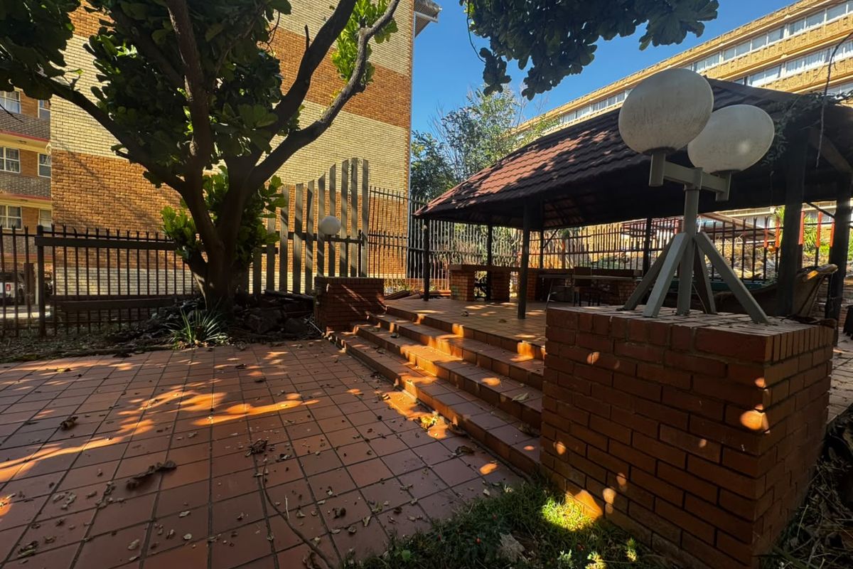 A communal outdoor area featuring a brick-paved patio, a covered gazebo with a built-in braai, and established trees within a fenced apartment complex.