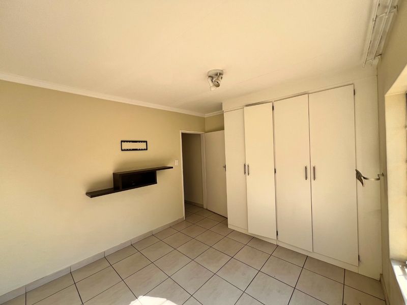 A clean, empty bedroom featuring neutral tiled flooring, built-in white cupboards, and a wall-mounted shelf, offering a blank canvas for personalization.