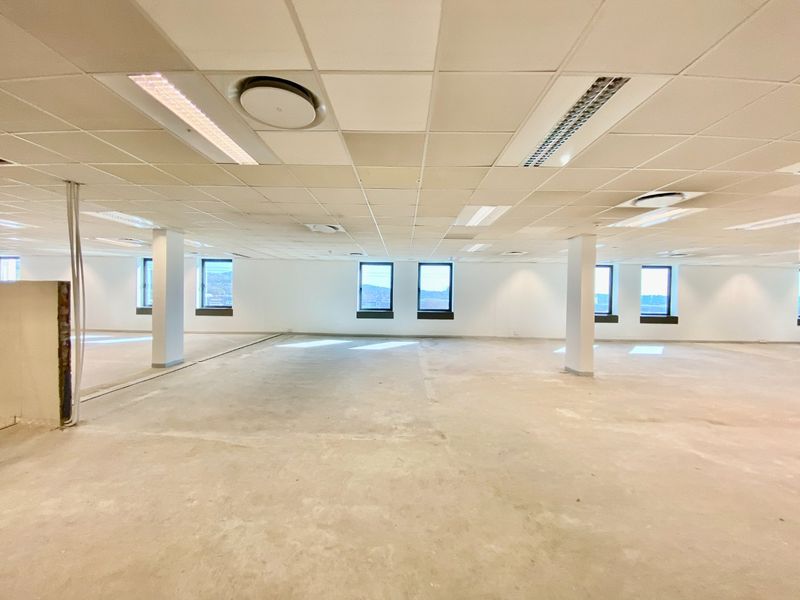 An expansive, open-plan commercial office space featuring a suspended ceiling, recessed lighting, structural columns, and large windows with views.