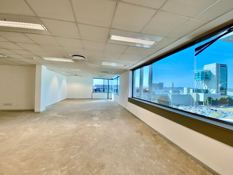 An open-plan commercial office space featuring a suspended ceiling, recessed lighting, and large windows with expansive city views, currently in a shell condition.