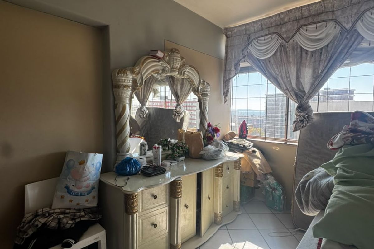 A bedroom featuring tiled flooring, a ceiling fan, and a built-in cupboard, with large windows offering an urban view.