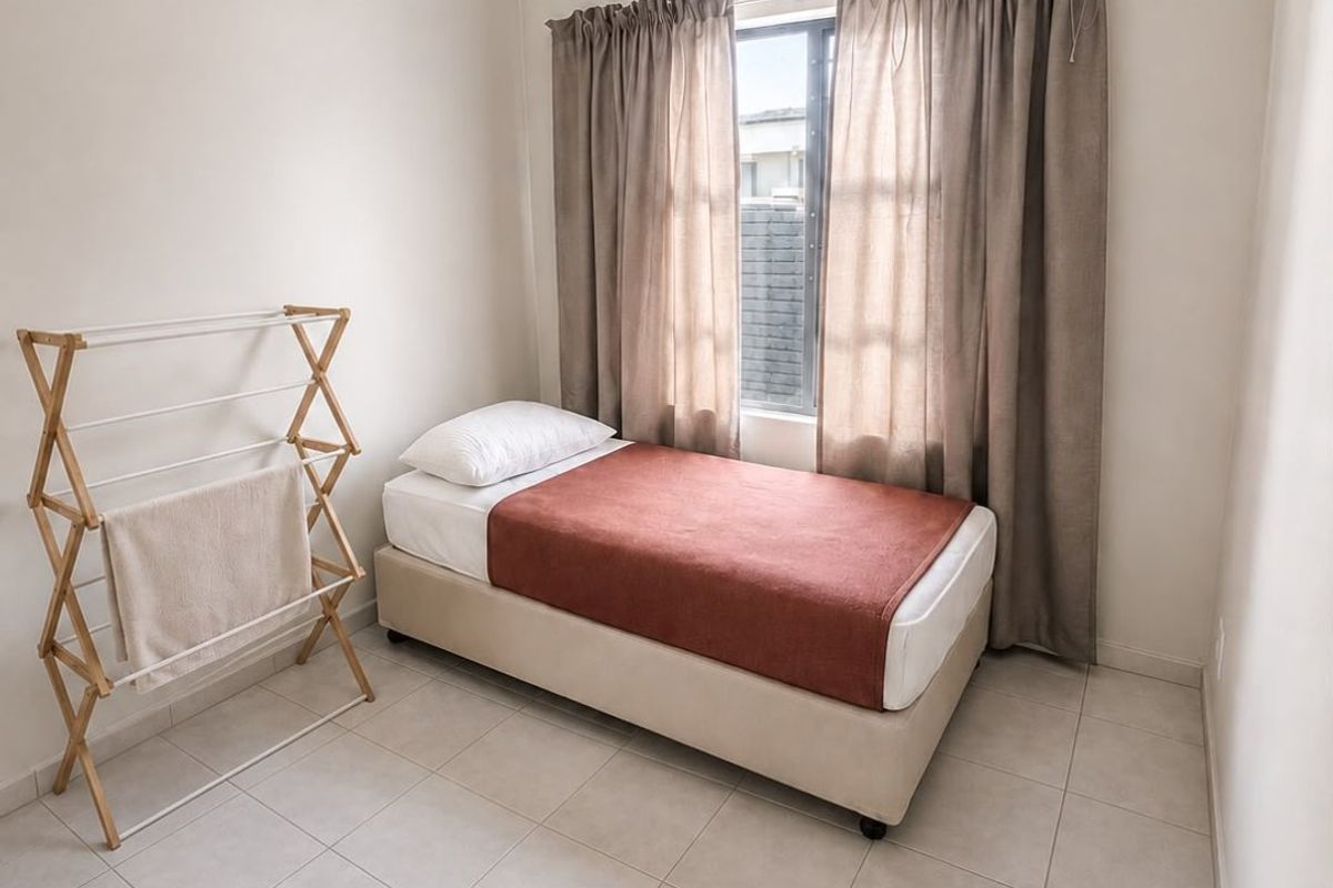 A compact bedroom featuring neutral tiled flooring, a single window with curtains, and a ceiling light fixture.