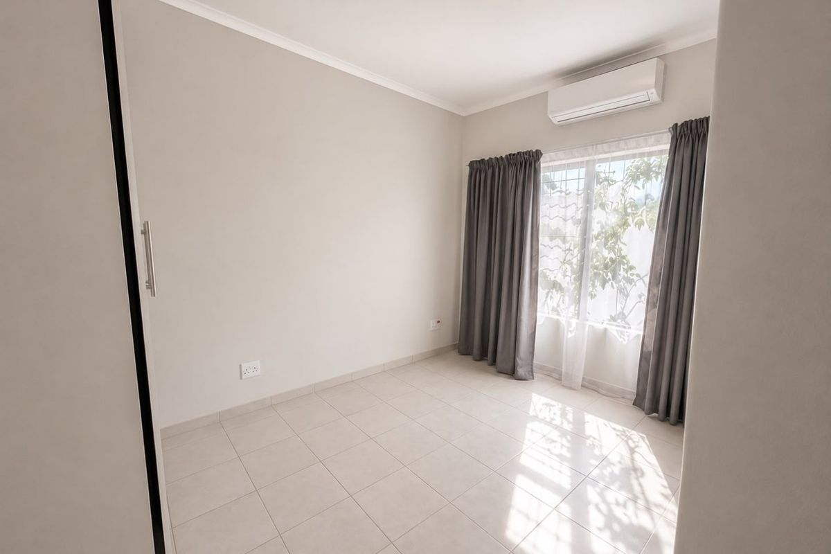A bright, empty bedroom featuring neutral tiled flooring, a built-in wardrobe, and a wall-mounted split-system air conditioner.
