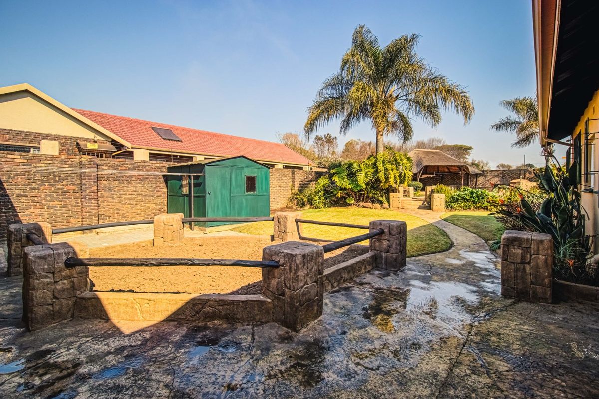 A spacious rear garden featuring a paved walkway, a sand pit with stone-look fencing, a garden shed, and a distant thatched lapa area.