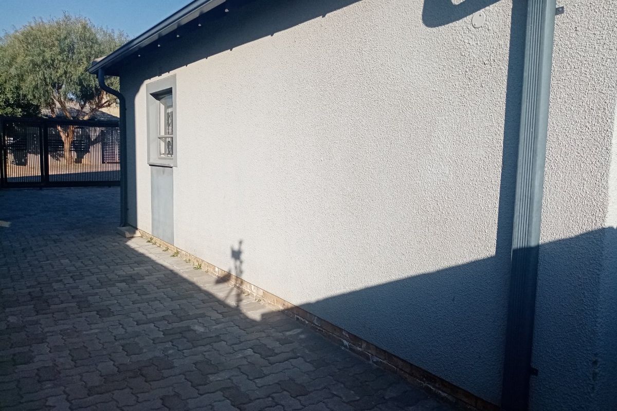 Side view of the property showing a textured plaster wall, downpipe, and paved driveway leading to a security gate.