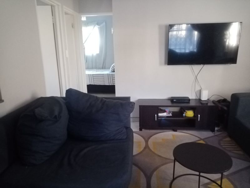 A living room area featuring tiled flooring and a wall-mounted television, with a view leading into an adjacent bedroom.