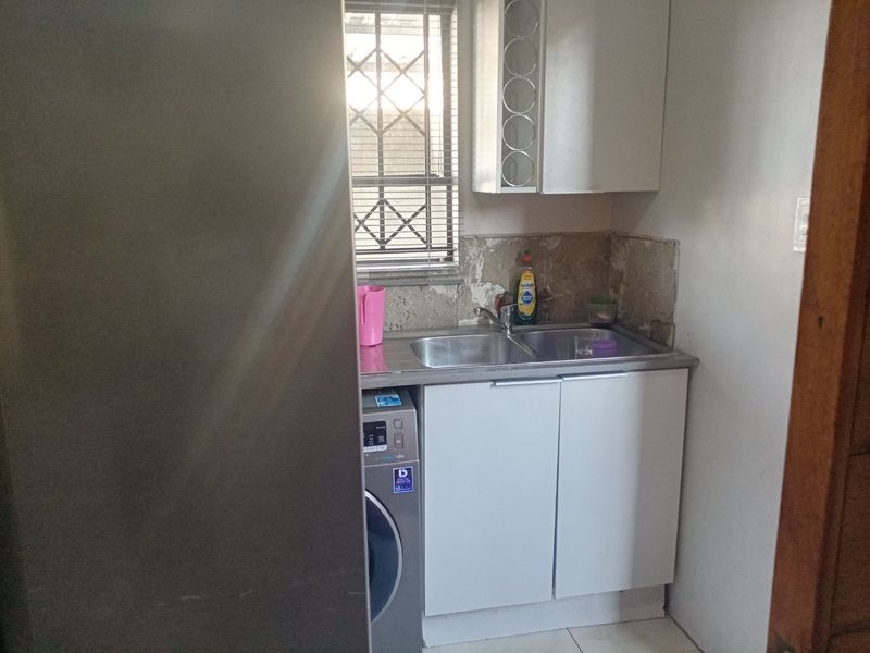 A compact scullery area featuring a stainless steel double sink, built-in cabinetry, and a window with security bars, requiring wall finish repairs.