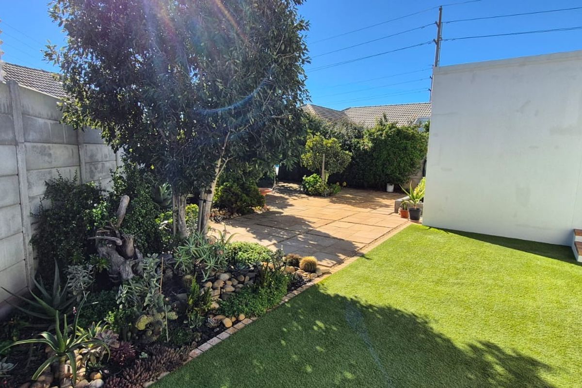 A well-maintained rear garden featuring a paved patio, artificial lawn, and established landscaping enclosed by a precast concrete boundary wall with electric fencing.