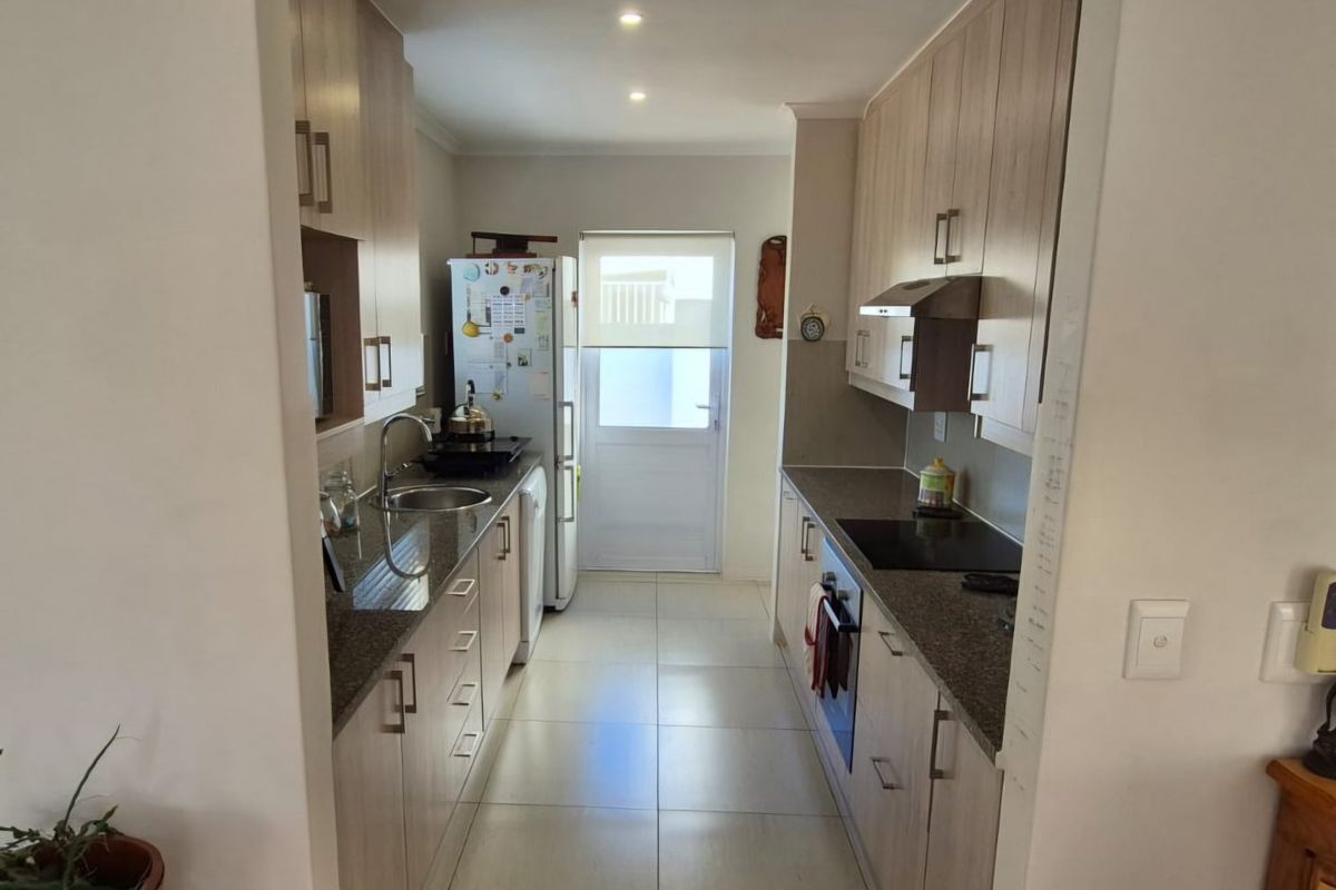 A functional galley-style kitchen featuring light wood-tone cabinetry, granite countertops, integrated appliances, and tiled flooring with direct access to the exterior.