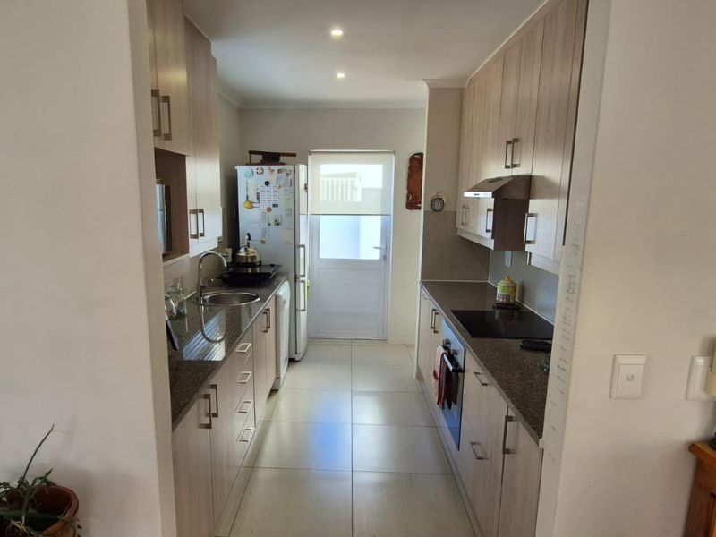 A functional galley-style kitchen featuring light wood-tone cabinetry, granite countertops, integrated appliances, and tiled flooring with direct access to the exterior.