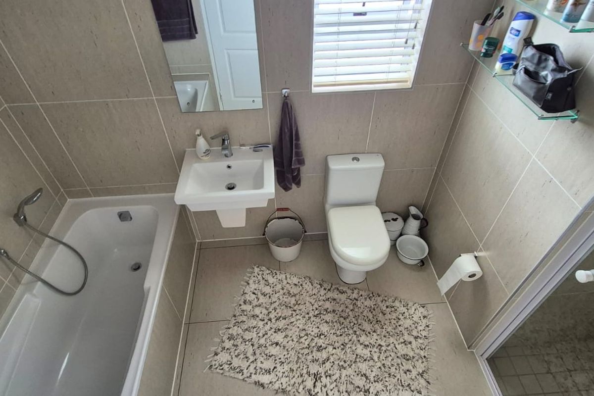 A compact, modern bathroom featuring a bathtub, wall-mounted basin, toilet, and neutral floor-to-ceiling tiling with natural light from a window.