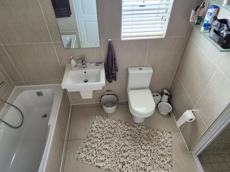 A compact, modern bathroom featuring a bathtub, wall-mounted basin, toilet, and neutral floor-to-ceiling tiling with natural light from a window.