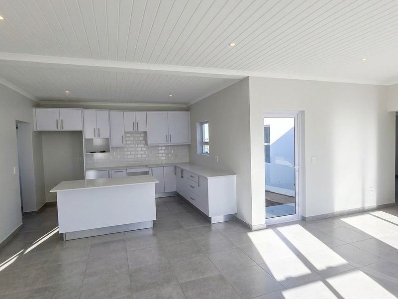 A bright, modern open-plan kitchen featuring white cabinetry, a central island, subway tile backsplash, and neutral tiled flooring, leading to an outdoor area.