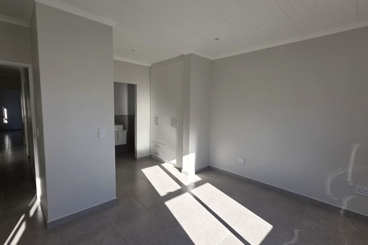 A bright, empty bedroom featuring neutral tiled flooring, built-in cupboards, and a clean tongue and groove ceiling, with an adjoining en-suite bathroom area.