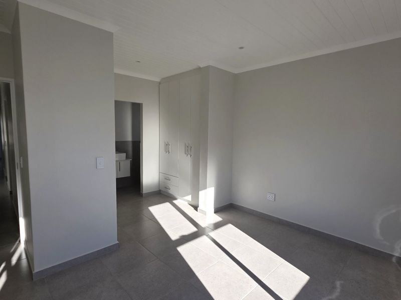 A bright, empty bedroom featuring neutral tiled flooring, built-in cupboards, and a clean tongue and groove ceiling, with an adjoining en-suite bathroom area.