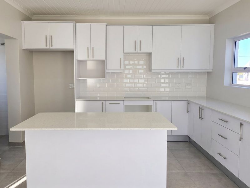 A modern, bright kitchen featuring white cabinetry, stone countertops, a subway tile backsplash, and a central island, finished with neutral floor tiles.