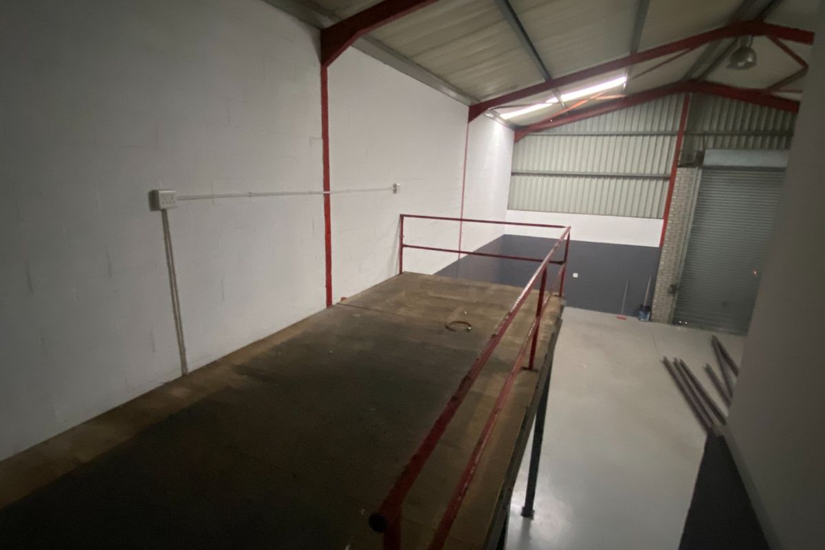 Industrial warehouse space featuring a mezzanine level with safety railing, concrete flooring, and a large roller shutter door for vehicle access.