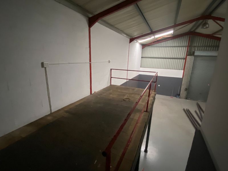 Industrial warehouse space featuring a mezzanine level with safety railing, concrete flooring, and a large roller shutter door for vehicle access.
