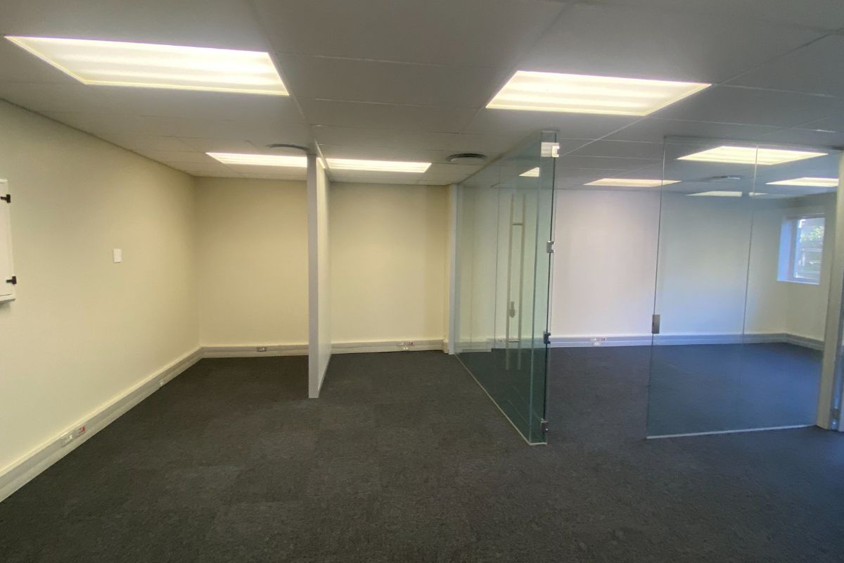 An empty commercial office space featuring glass partition walls, carpeted flooring, and a suspended ceiling with recessed lighting.