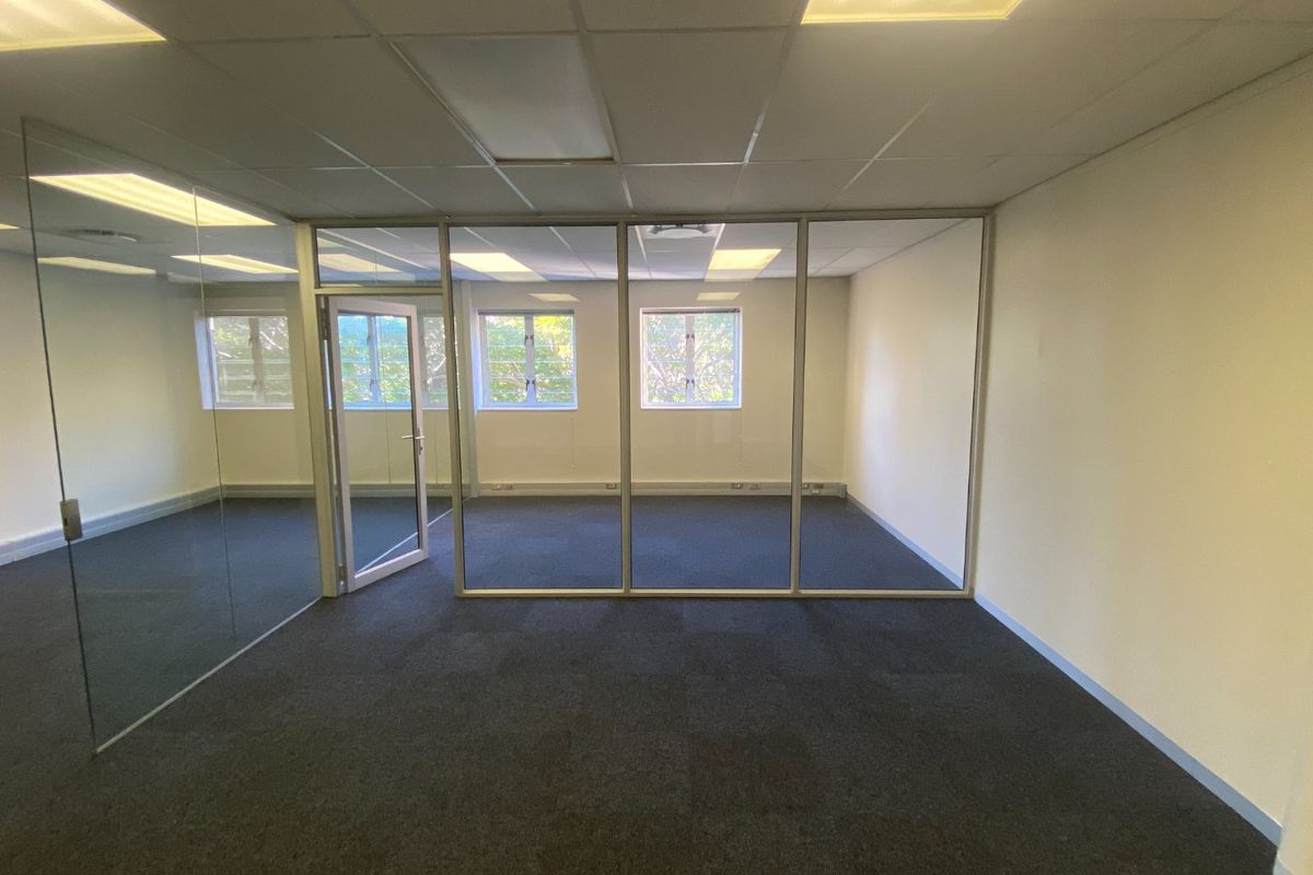 A vacant commercial office space featuring glass partition walls, carpeted flooring, and a suspended ceiling with recessed lighting.
