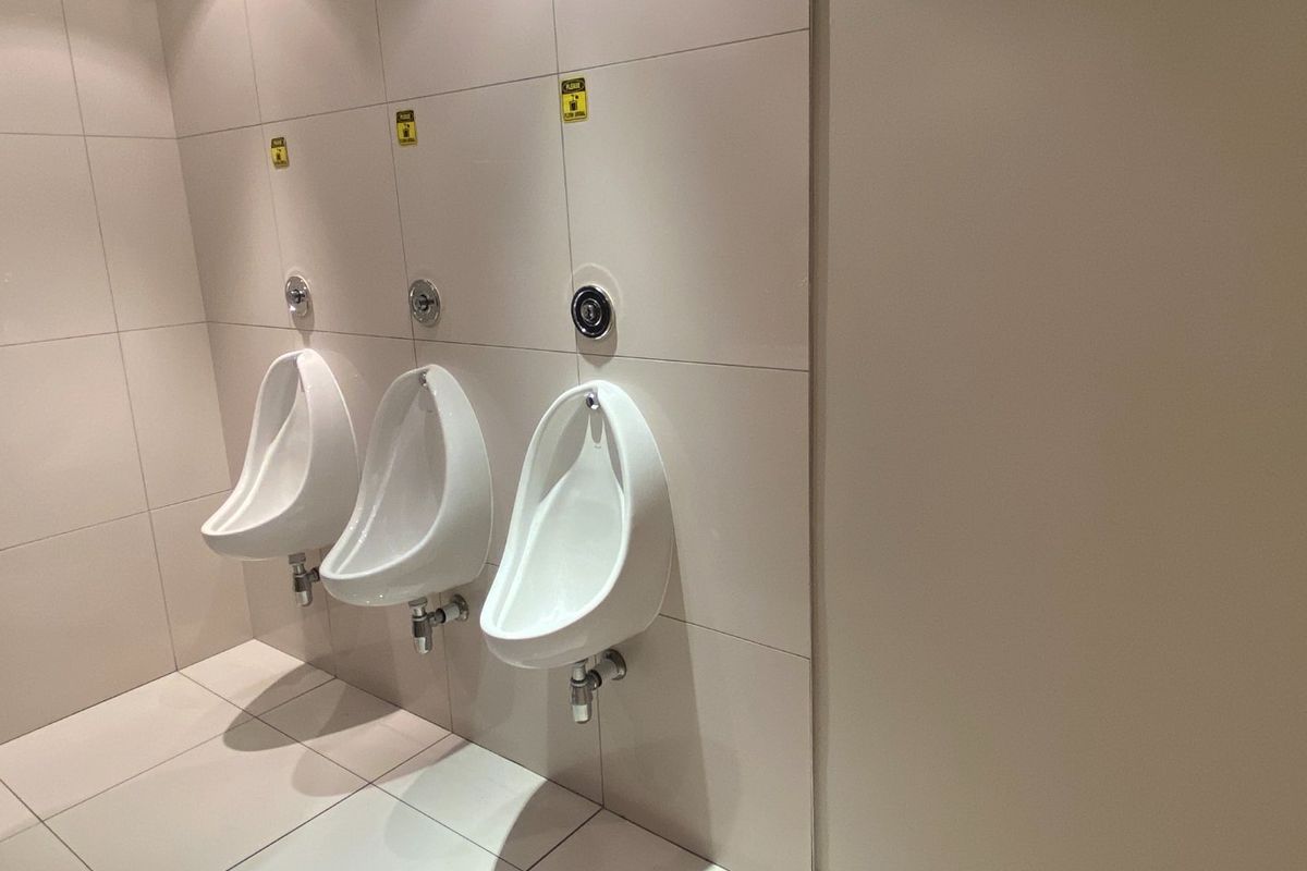 A clean, modern commercial restroom featuring three wall-mounted ceramic urinals with integrated flush valves and neutral tiled finishes.