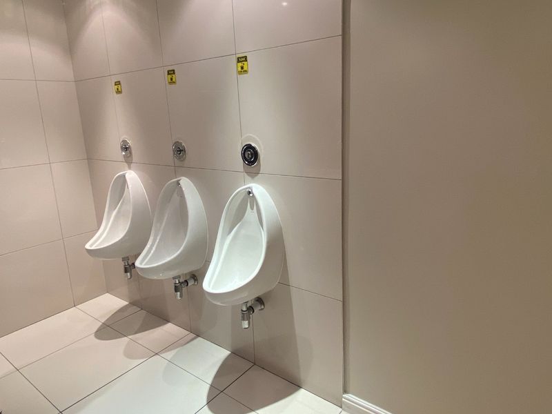 A clean, modern commercial restroom featuring three wall-mounted ceramic urinals with integrated flush valves and neutral tiled finishes.