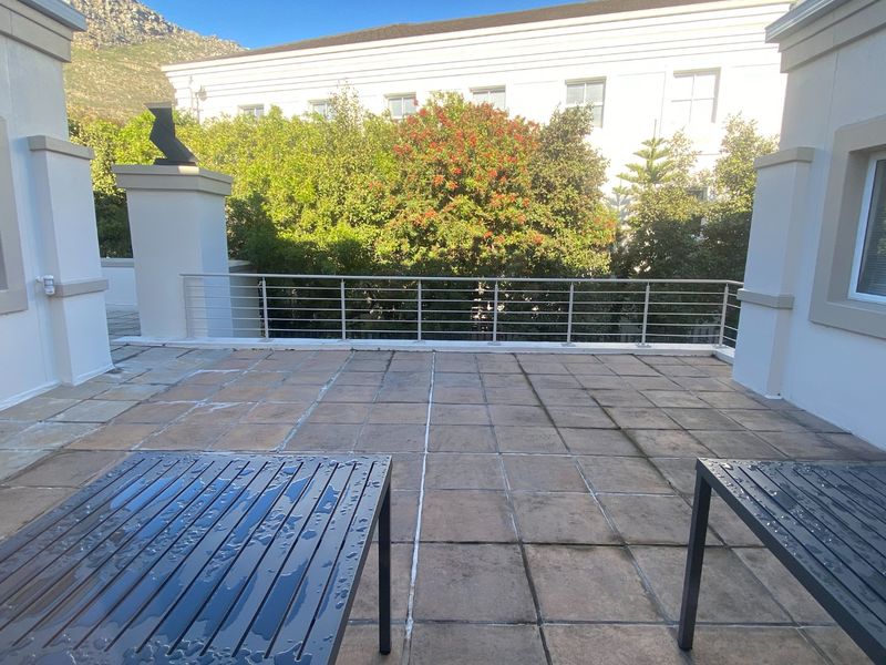 An open-air balcony featuring tiled flooring, a metal balustrade, and masonry pillars, offering a clear view of the surrounding greenery and building exterior.