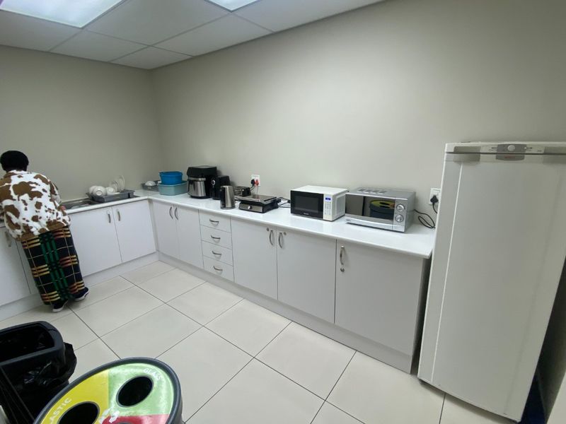 A functional commercial kitchenette featuring white built-in cabinetry, laminate countertops, a sink, and tiled flooring with a drop ceiling.