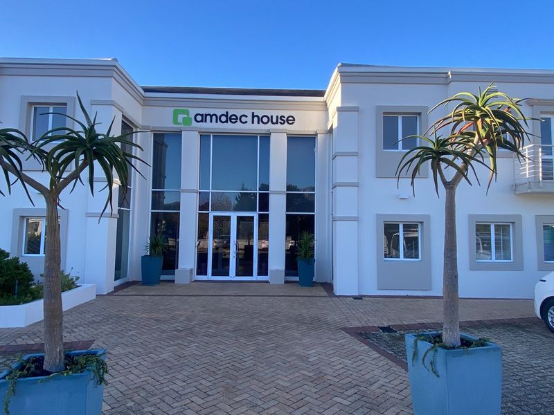 Front elevation of Amdec House, a modern commercial building featuring a prominent glass entrance, paved driveway, and well-maintained white facade.
