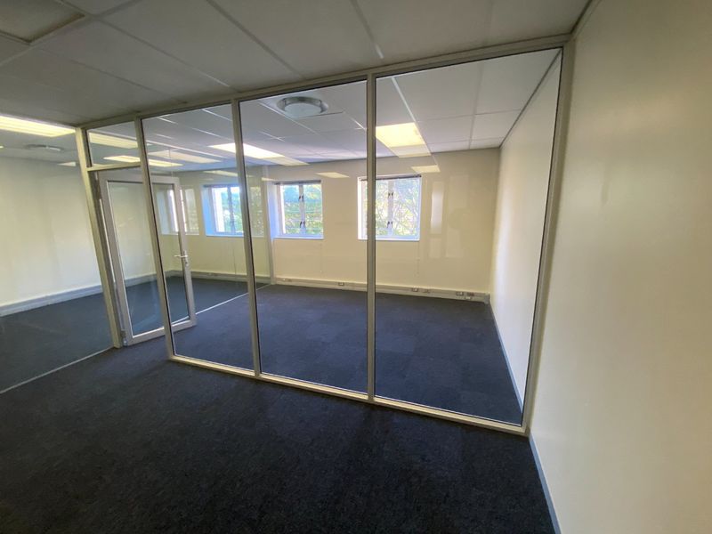 A vacant commercial office space featuring glass partitioning, carpeted flooring, and a suspended ceiling with recessed lighting, offering a clean and professional work environment.