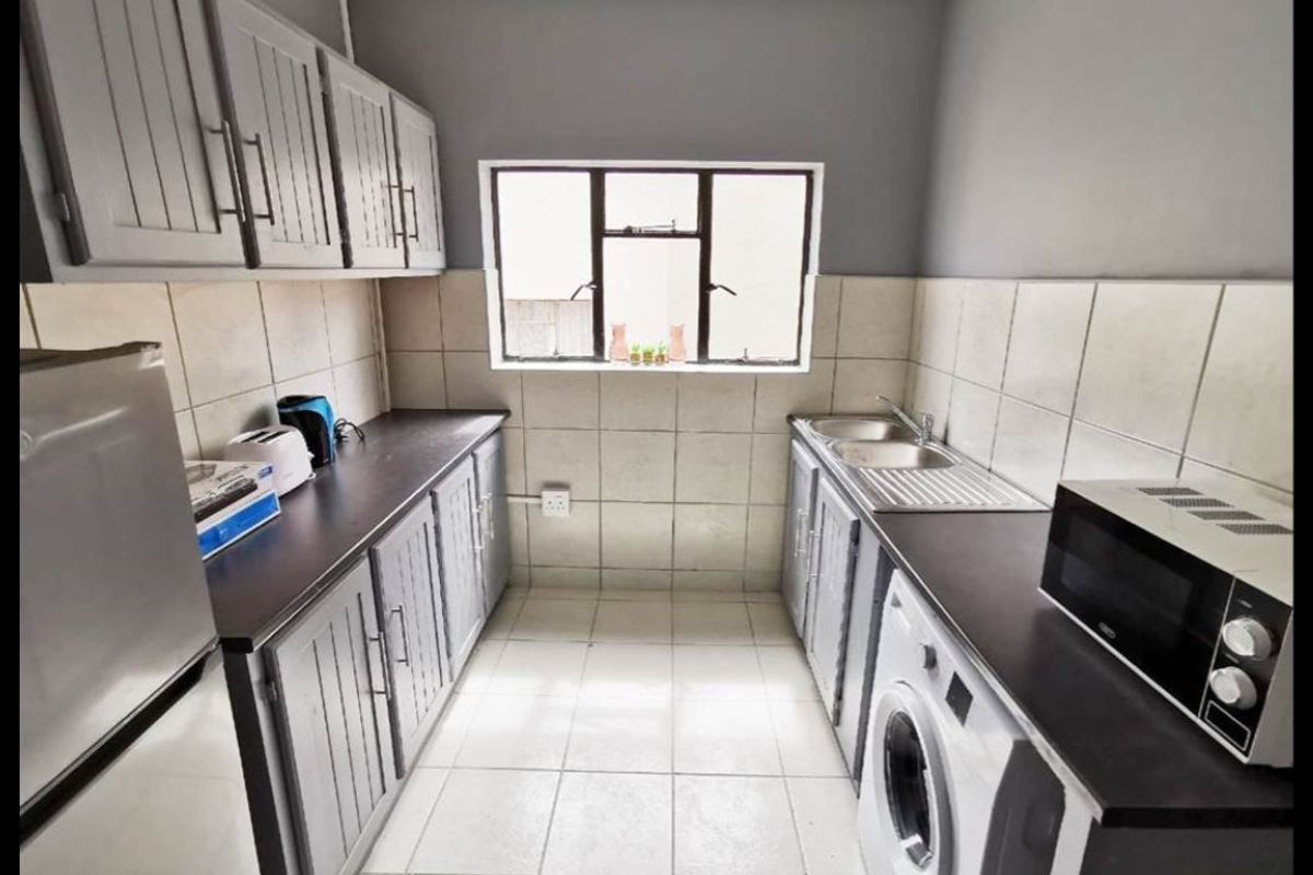 A functional galley-style kitchen featuring grey cabinetry, dark laminate countertops, tiled backsplash, and integrated space for laundry appliances.